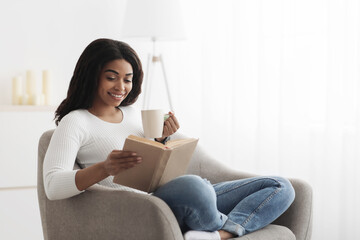 Fototapeta premium Free time to relax. Calm black woman relaxing on comfortable chair with paper book and holding cup of hot coffee. Happy lady taking break from work, copy space