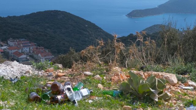Environmental pollution. Pile of discarded glass bottles littering a grassy hill, creating a stark contrast with the stunning panoramic view of a seaside village, blue ocean, and green mountains