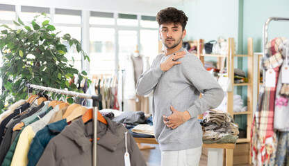 Young man in twenties trying on warm light grey sweatshirt (grey knitted sweater) in local clothing store, checking fit and testing comfort before buying while shopping for casual wardrobe items