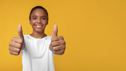 Cheerful teen african american girl in white t-shirt rejoices to success and shows thumbs up, close...