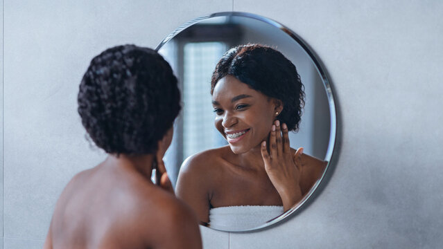 A woman stands in front of a round mirror smiling at her reflection while applying a skin care product. She is in a bathroom with light colors and simple decor.