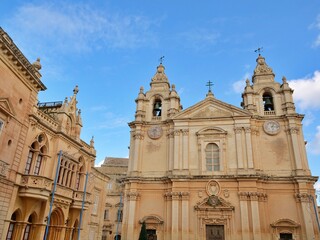 Fototapeta premium St. Paul's Cathedral in Mdina, Malta, features twin bell towers and ornate baroque limestone architecture under a blue sky with clouds, photographed from a low angle.