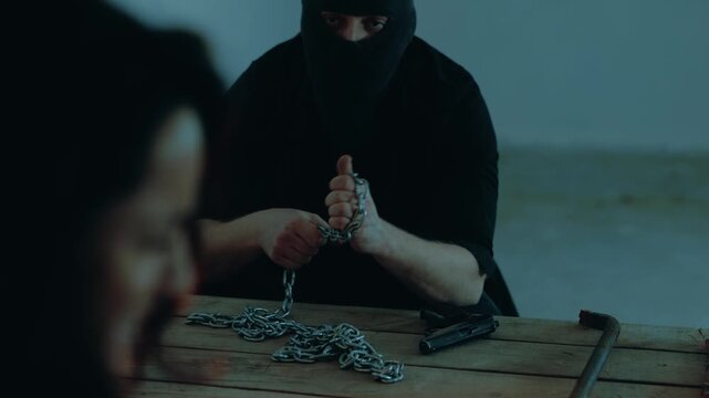 Male kidnapper in a black balaclava winding a chain around his arm and looking at the hostage. Frightened woman closing her eyes. Kidnaping, crime, violence concept. Slow motion