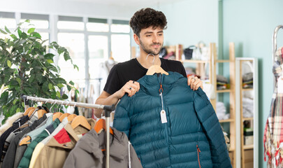 Young man in twenties updating wardrobe, carefully choosing winter puffer jacket in local clothing...