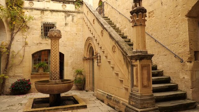 Palazzo Falson Historic House Museum courtyard in Mdina, Malta, featuring a stone fountain, stairs with a lion sculpture on the newel, and traditional limestone architecture under a bright, sunny sky.