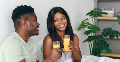 A man and a woman sit on a bed in a bright room. They share smiles and hold glasses of orange juice. Green plants and shelves are in the background. Morning light fills the space.