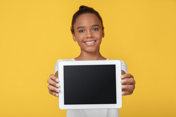 Smiling pretty teen african american girl showing tablet with blank screen, isolated on yellow...