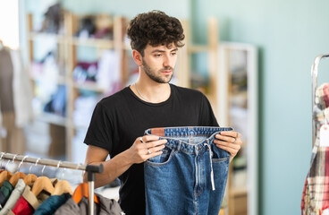 Regular customer guy client pre-examine fabric and style of product, is busy choosing jeans trousers. Client enjoys shopping for clothes at store browsing through colorful on display racks