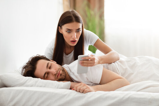 Jealousy Problem. Shocked Girlfriend Reading Messages On Cheating Husband's Phone Suspecting Infidelity In Bedroom At Home. Jealous Girlfriend Checking Boyfriend's Smartphone. Selective Focus