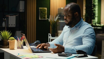 Black businessman sighing and checking messages on mobile phone, feeling drained and frowning after being distracted by a notification in modern office. Workplace challenges at night.