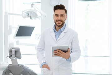 Fototapeta premium Attractive young bearded man dentist keeping beautiful smile on his face and holding digital tablet over cabinet background, empty space. Online application at dentist clinic concept