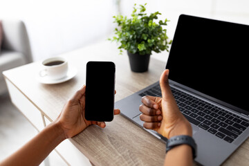 Unrecognizable black woman with cellphone and laptop recommending new mobile shopping app or website, showing thumb up gesture at home, mockup for design. Modern technologies in everyday life