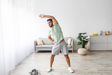 Full length of athletic young Arab man bending aside, doing exercises at home, copy space....