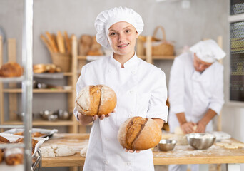 Enthusiastic young female baker apprentice presenting fresh baked loaves of bread with satisfied smile, standing in warm inviting bakery filled with variety of baked goods..