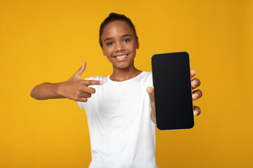 Cheerful teen afro american female pupil in white t-shirt points finger at smartphone with blank...