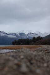 A scenic view of Cerro Tronador snow-covered mountains and a lake with a pebbly shore and dry reeds under heavy grey clouds during early winter in Patagonia, Argentina. Low angle shot, no people.