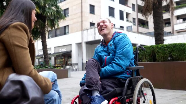 Young man with a physical disability in a wheelchair enjoying a conversation with a female friend in a city square, smiling and laughing together, representing social inclusion and happiness