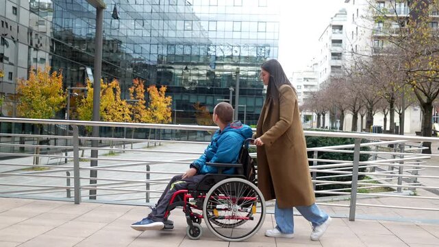Young disabled man in a wheelchair and his female caregiver strolling along a modern city street, enjoying a moment together with urban architecture in the background on an autumn day