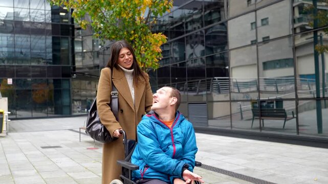Young caregiver assisting a man with cerebral palsy in a wheelchair, strolling and talking together on a city street in front of a modern glass building, representing inclusion and support