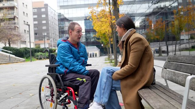 Young man with a disability sitting in a wheelchair having an engaging conversation with a female friend on a city park bench, sharing a moment of connection and support in an urban environment