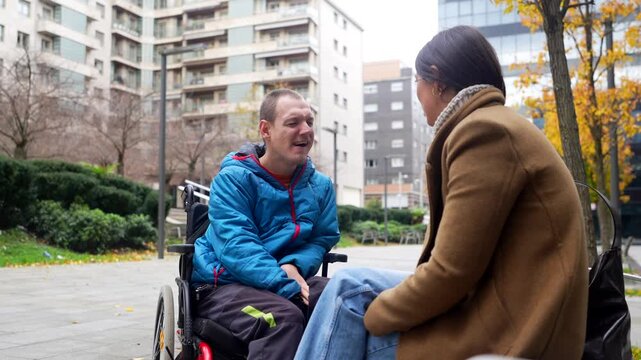 Young disabled man in a wheelchair having an animated and happy conversation with a female caregiver or friend in an urban park, showcasing supportive relationships and positive social interaction