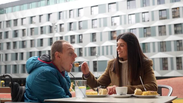 Young woman caring for a man with a disability, sitting together at a cafe table outdoors where she helps him by feeding him a piece of cake while they chat and spend quality time