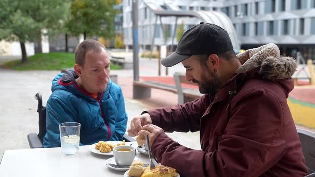 Caring friend or social worker helping a young man with a disability, feeding him lunch at an outdoor restaurant table, showing support, inclusion, and genuine human connection