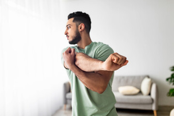 Athletic young Arab guy stretching arm muscles, doing warmup exercises at home, selective focus....