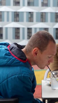 Young man with a physical disability in a wheelchair drinking from a straw and talking with his girlfriend at an outdoor cafe table, enjoying a moment of connection and normalcy together