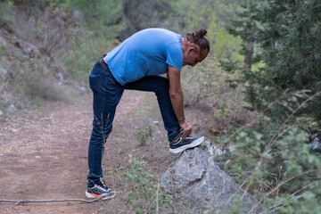 Man Tying Shoelaces During a Forest Walk. Hiker Adjusting Shoes on a Nature Trail. Outdoor Walk Pause to Tie Shoe Laces. Preparing for Hiking in the Forest.