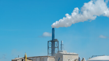 Industrial chimney emitting thick white steam against a clear blue sky. Industrial plant architecture with steel structures and factory buildings
