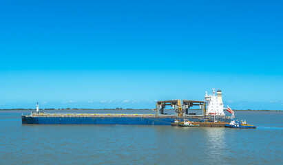 Large blue bulk carrier ship with heavy-duty cranes anchored in calm coastal waters, assisted by tugboats. Industrial marine