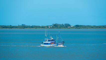 Small blue and white fishing trawler cruising through calm blue coastal waters on a sunny day with a green shoreline in the background