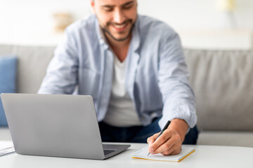 Obraz premium Male freelancer writing in notepad and using laptop, taking notes at workplace, using computer, noting information from internet, selective focus on male hand, closeup