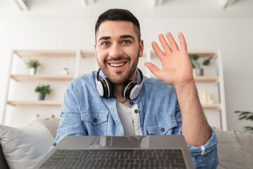 Greeting Concept. Portrait of excited young man waving hello with hand looking at camera, using laptop at home office. Cheerful guy saying hi or goodbye during video call, webcam view © Prostock-studio
