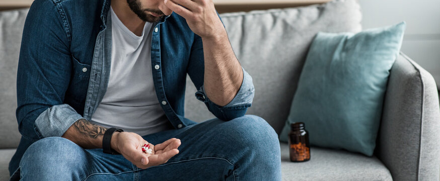 Addiction to painkillers, drug addiction, overdose, suicide and antidepressants. Sad unhappy despair adult european male hold on palm a lot of different pills sits on couch with jar, cropped, panorama