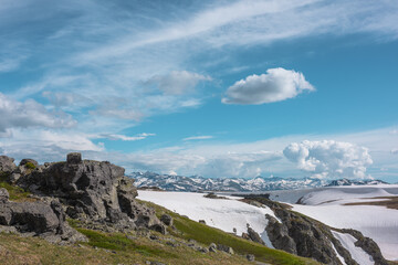 Scenic view to sunlit grassy hillside with rocky outcrops against snowfields on rocks and big snowbound rock mountain range far away under clouds in blue sky. High snow mountains in changeable weather © Daniil