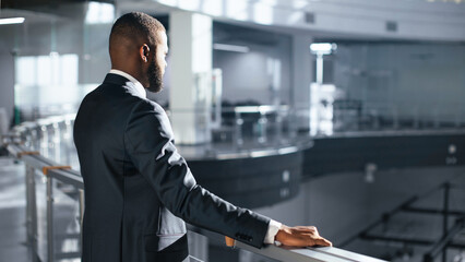 A businessman in a suit stands on a balcony, observing a spacious office area below. Natural light...
