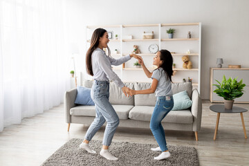 Joyful asian mom and daughter dancing to music, holding hands and enjoying favorite song together, having fun in living room interior at home. Full length shot