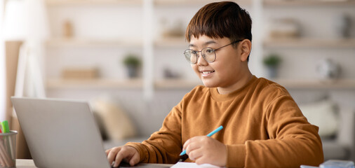 A young boy sits at a table working on his laptop. He takes notes with a blue pen and smiles as he engages with his schoolwork in a cozy study area during the day. © Prostock-studio