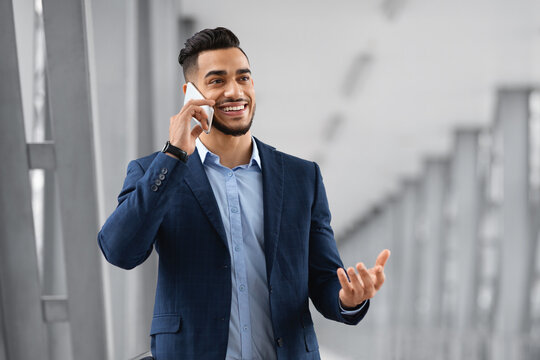 Handsome Arab Entrepreneur Talking On Mobile Phone While Waiting For Flight Departure At Airport, Smiling Middle Eastern Businessman In Suit Standing At Terminal And Enjoying Cellphone Conversation