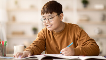 A young boy sits at a table with a notebook and colored pens. He smiles as he writes down notes. A cup of drink is placed nearby. The setting is a home with bookshelves in the background. © Prostock-studio