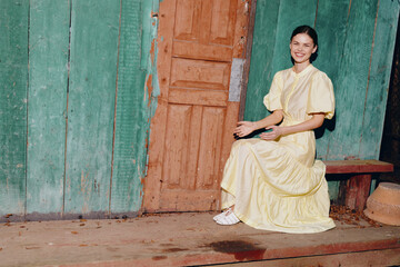 Woman in a pale yellow dress sits on a wooden bench beside a weathered doorway, against a turquoise...
