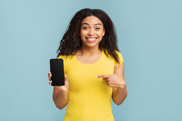 Naklejka premium Trendy mobile phone. Happy black woman pointing at smartphone with blank screen over blue background, cellphone with space for mobile app design mockup