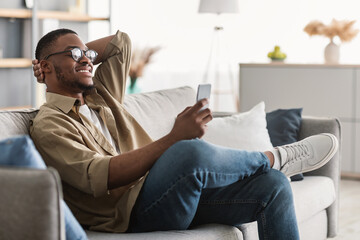 Fototapeta premium New Mobile App. Cheerful Black Man Using Cellphone Wearing Eyeglasses, Texting And Browsing Internet Sitting On Couch At Home. Phone User Using Application Indoors. Side View