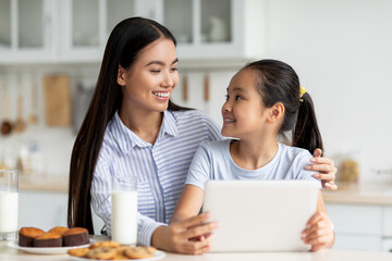 Loving asian mother and her daughter using digital tablet, looking for new recipes while sitting at kitchen table and enjoying cookies with milk. Young mom and child spending time together