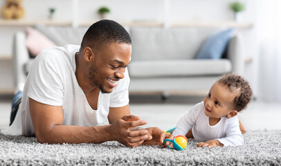 Plakat Father-infant bonding. Portrait of happy young black dad playing with baby son at home, cute little african american toddler child relaxing with daddy on floor in living room, spending time together