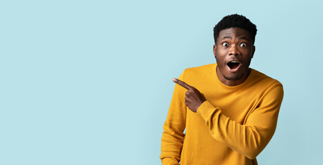 Emotional millennial black guy in yellow sweater pointing at empty space for advertisement over...