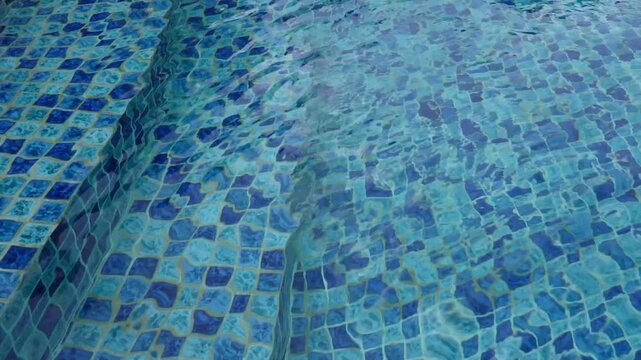 Footage of clear blue swimming pool water with gentle ripples flowing over mosaic tile steps. Relaxing abstract background suitable for summer, resort, vacation, spa, and luxury leisure concepts.
