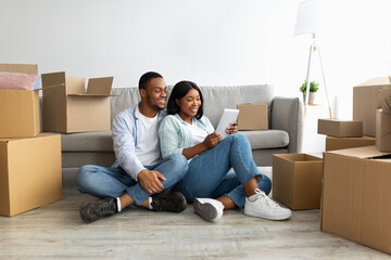 Relocation concept. Happy black couple using tablet, searching new furniture while sitting among...